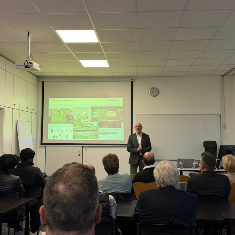 Man in a suit giving a presentation at the front of a classroom, with a slide deck projected on a screen behind him and an audience of professionals watching.