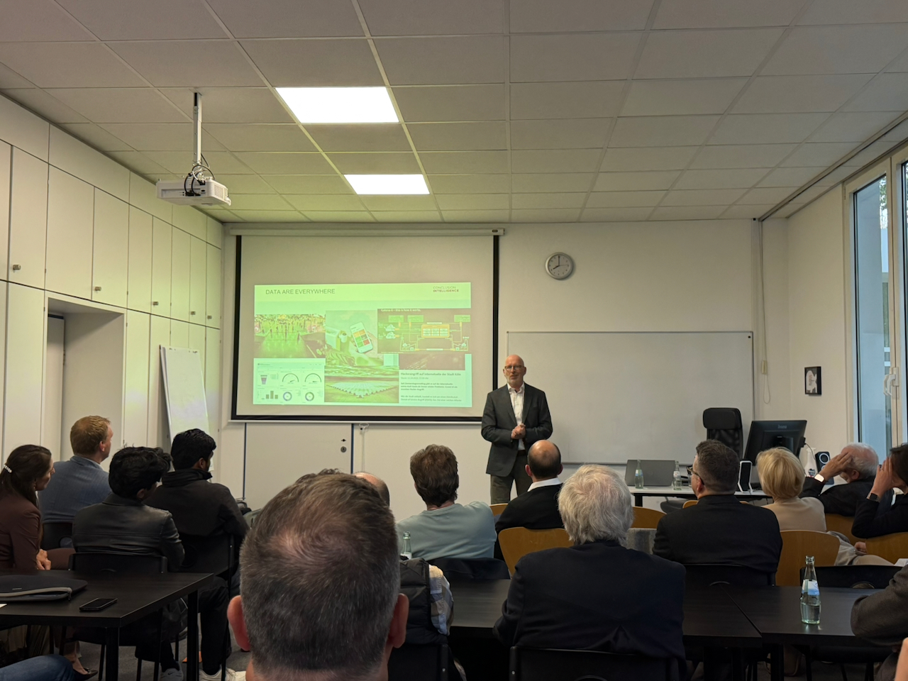 IMG_5138 Man in a suit giving a presentation at the front of a classroom, with a slide deck projected on a screen behind him and an audience of professionals watching.