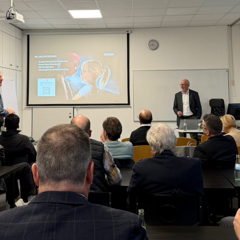 Conference room presentation with two men at the front and a projected slide about Spider-Man's tactics/text appears on screen.