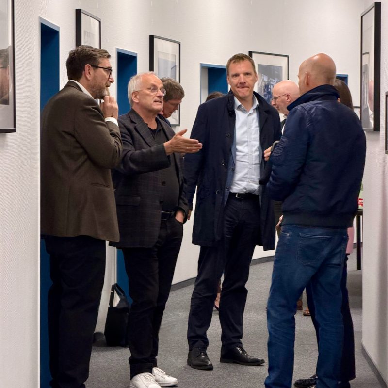 Group of men in business attire converse in a hallway lined with framed photos.