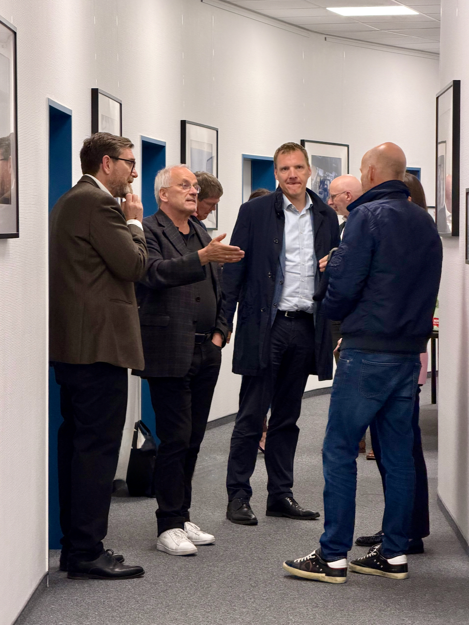 IMG_5150 Group of men in business attire converse in a hallway lined with framed photos.