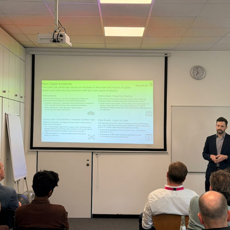 Presenter in a dark suit giving a lecture in a classroom with a projected slide titled 'Main Cyber Incidents' showing cyber risk content.