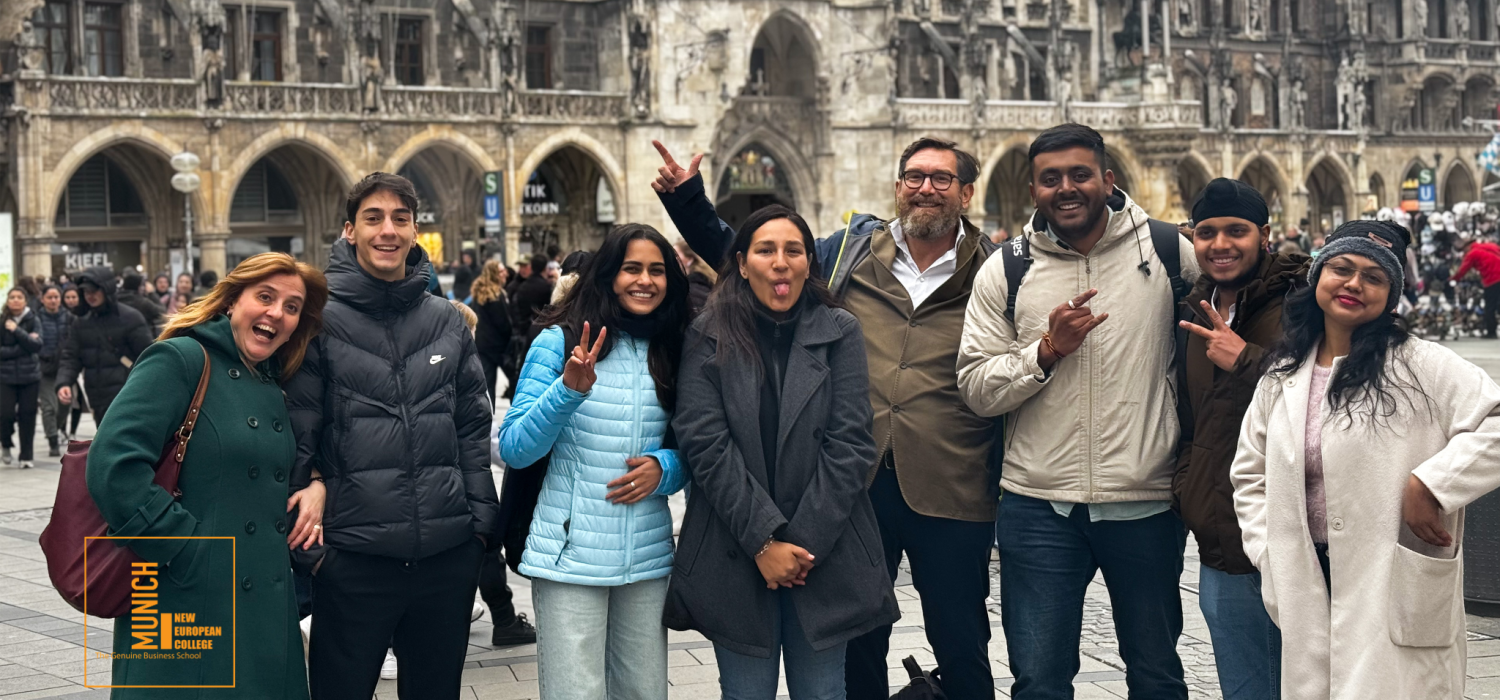 Group of diverse friends posing for a photo in a busy European square, with a historic stone building behind them and a bright orange MUNICH logo overlay in the corner.
