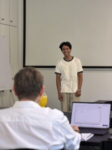Smiling young man giving a presentation at the front of a classroom, audience seated with laptops in view.