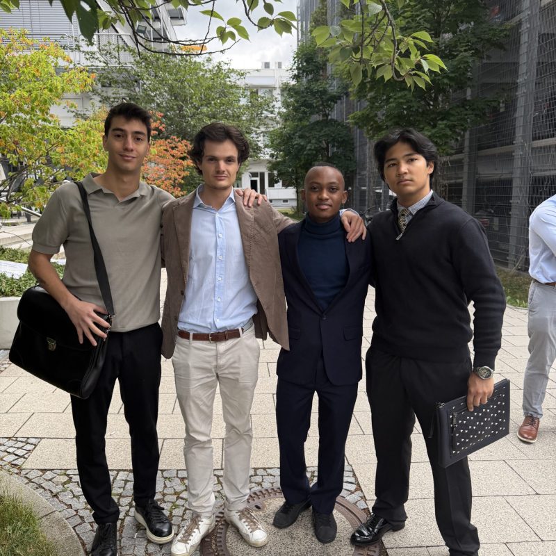 Four young men stand together outdoors in an urban courtyard, arms around each other's shoulders, smiling at the camera.