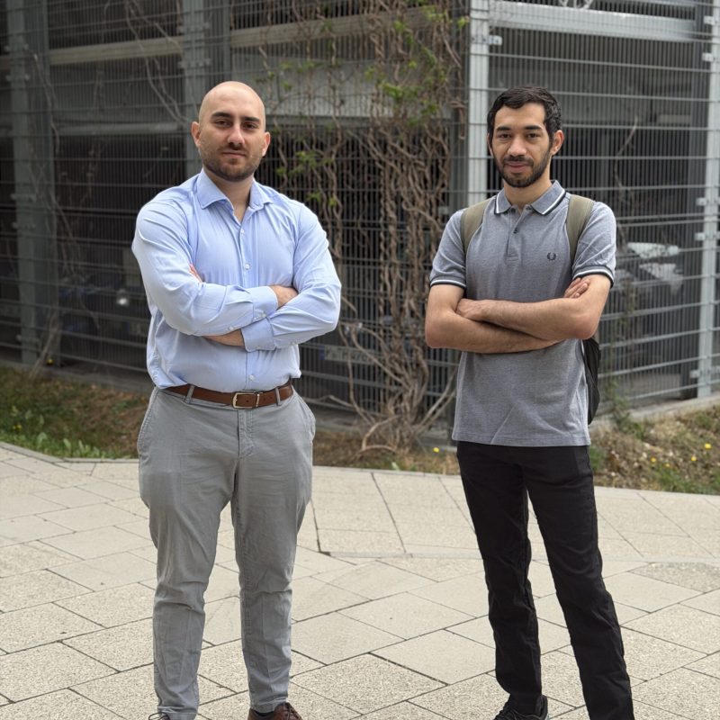 Two men stand with arms crossed in front of a metal fence; left man in light blue shirt and gray pants, right man in gray polo and backpack.