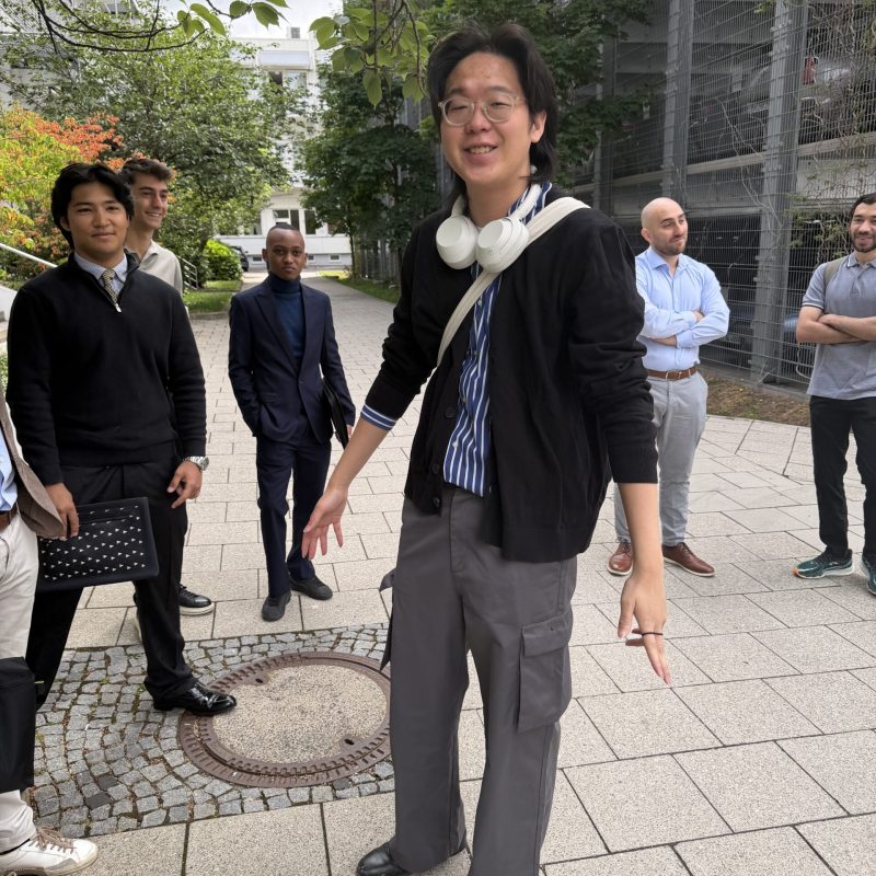 Smiling man with white headphones around his neck stands in front while colleagues chat outdoors in an urban courtyard.