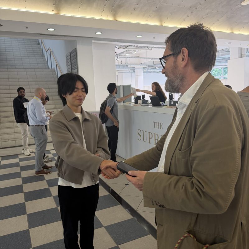 Two men shake hands in a modern lobby near a reception desk, with several people in the background and a staircase visible to the left.
