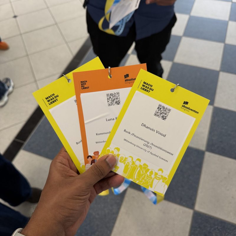 Close-up of a person holding bright yellow and orange booklets with QR codes, titled 'MACH WAS DU LIEBST' at a conference or event.