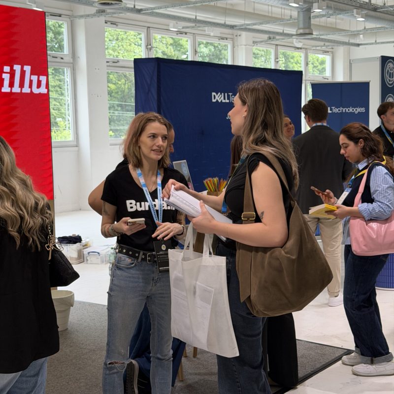 Two women stand at a business expo booth, exchanging papers and a tote bag amid blue and red display banners in a bright hall.