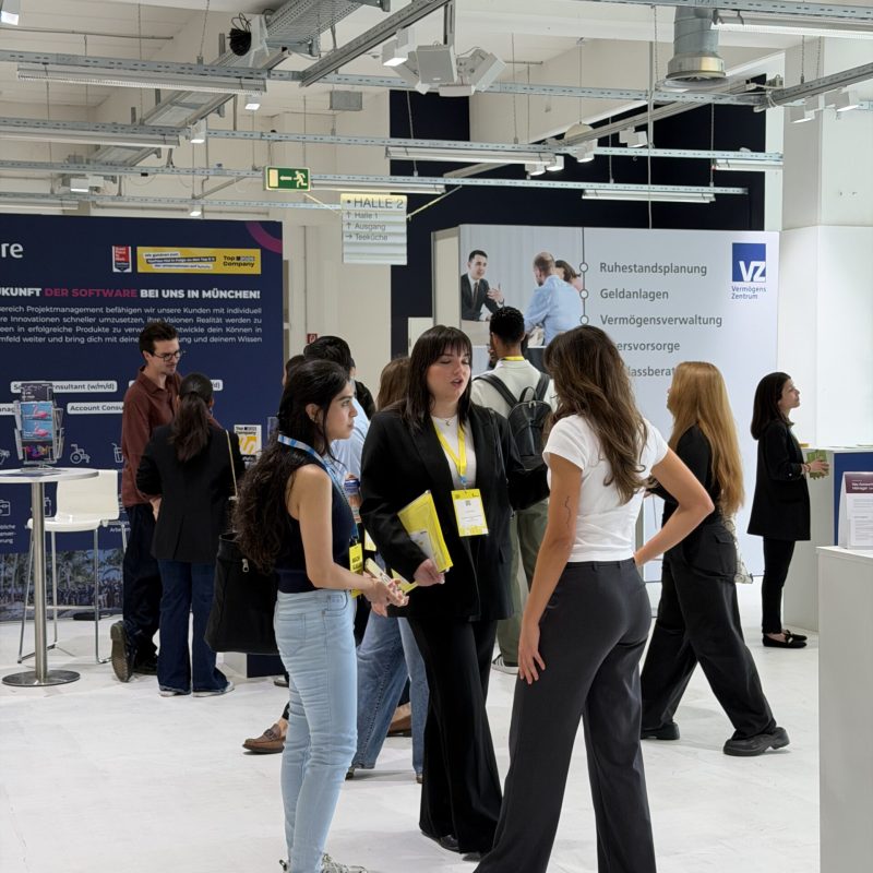 Crowd of convention attendees at an indoor exhibition, standing near booths with signs and banners under a high, industrial ceiling.