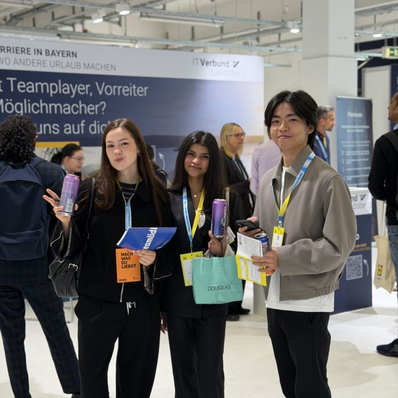 Three young adults at a trade show booth, smiling and posing with brochures and colorful bags.