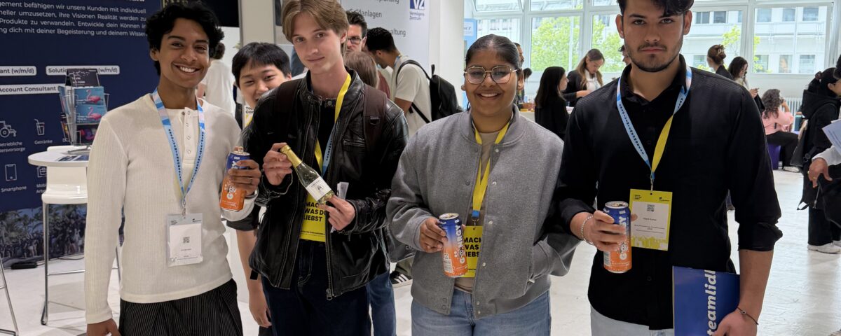 Five diverse young people at an indoor expo, holding beverages and wearing yellow lanyards, with booth banners in the background.