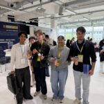 Five diverse young people at an indoor expo, holding beverages and wearing yellow lanyards, with booth banners in the background.