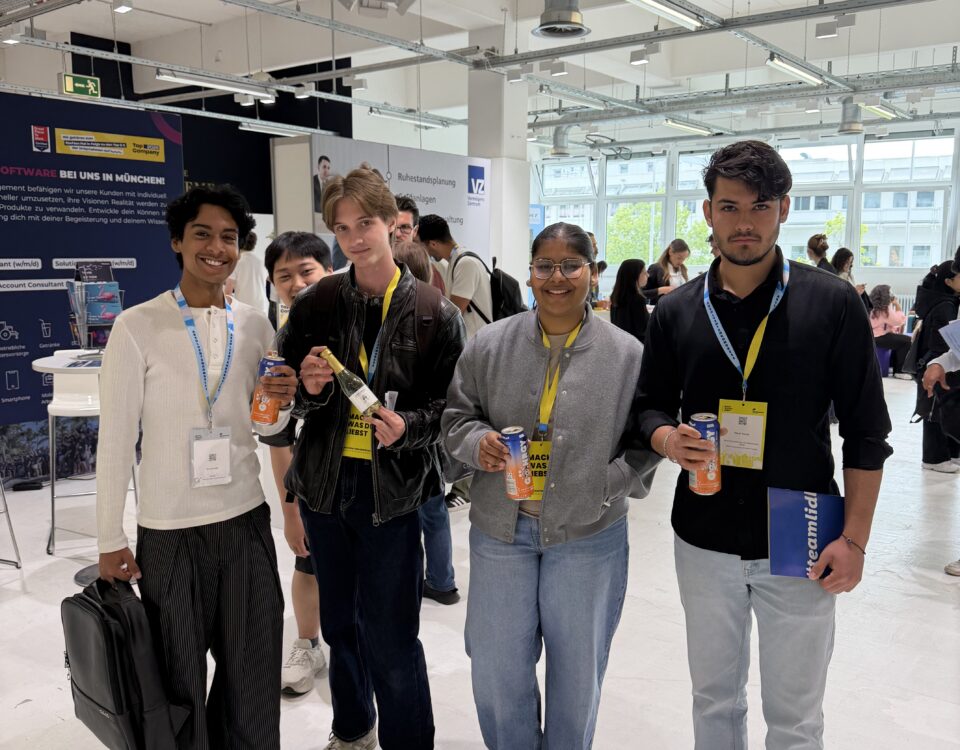 Five diverse young people at an indoor expo, holding beverages and wearing yellow lanyards, with booth banners in the background.