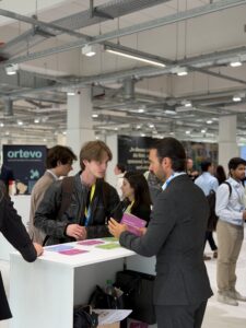 Two men in business casual attire talk at a white counter covered with colorful brochures in a crowded exhibition hall.