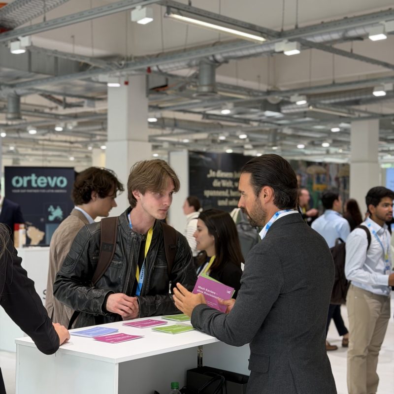 Two men in business casual attire talk at a white counter covered with colorful brochures in a crowded exhibition hall.