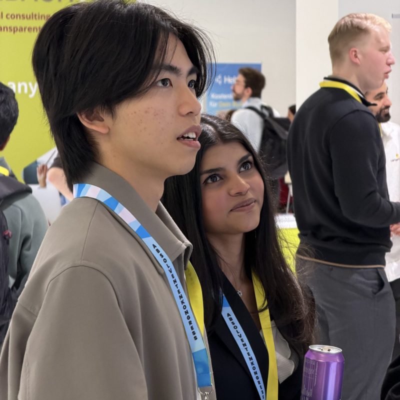 Two young attendees at a conference with lanyards; a man in a beige jacket in the foreground and a woman behind him, drinks in view and a yellow event banner in the background