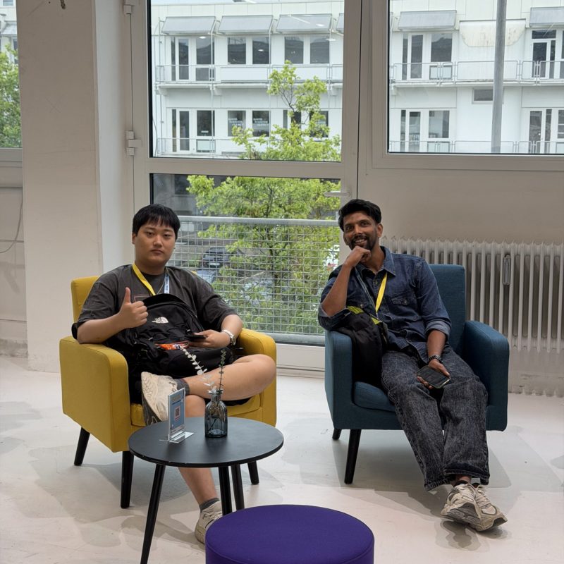 Two men wearing lanyards sit in bright yellow and blue chairs by large windows, one giving a thumbs up.