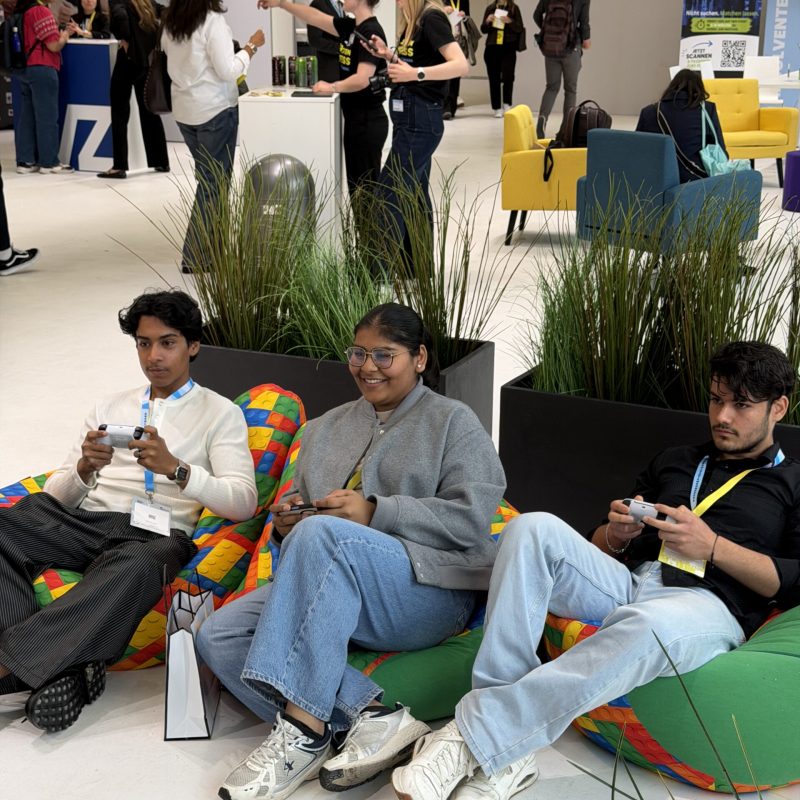 Three young people sit on colorful inflatable seats at a conference lounge, playing on handheld gaming devices with lanyards around their necks.