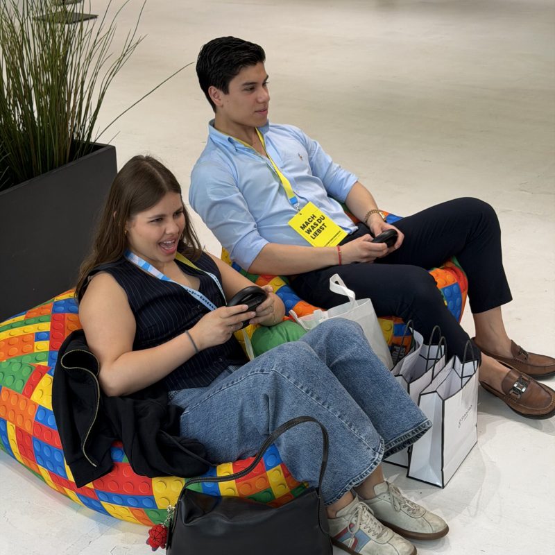 Two young adults relaxing on a bright Lego-pattern bean bag, looking at smartphones, with shopping bags nearby in a bright indoor space.