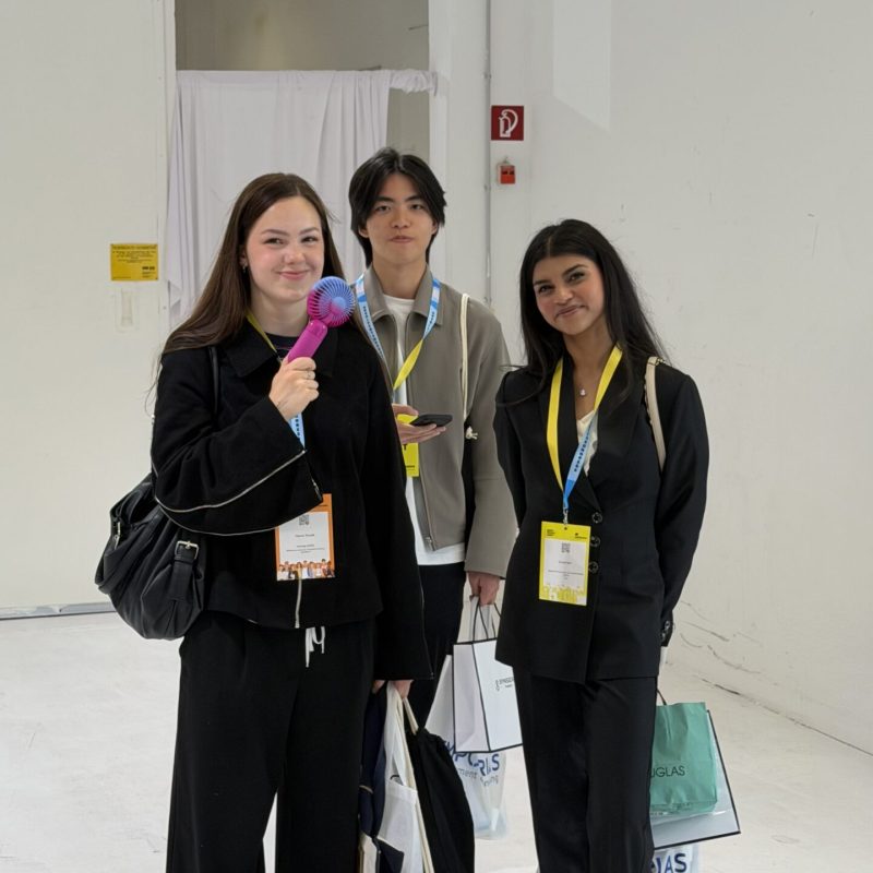 Three young adults with conference badges pose in a bright white studio; the person on the left holds a pink and blue handheld fan.