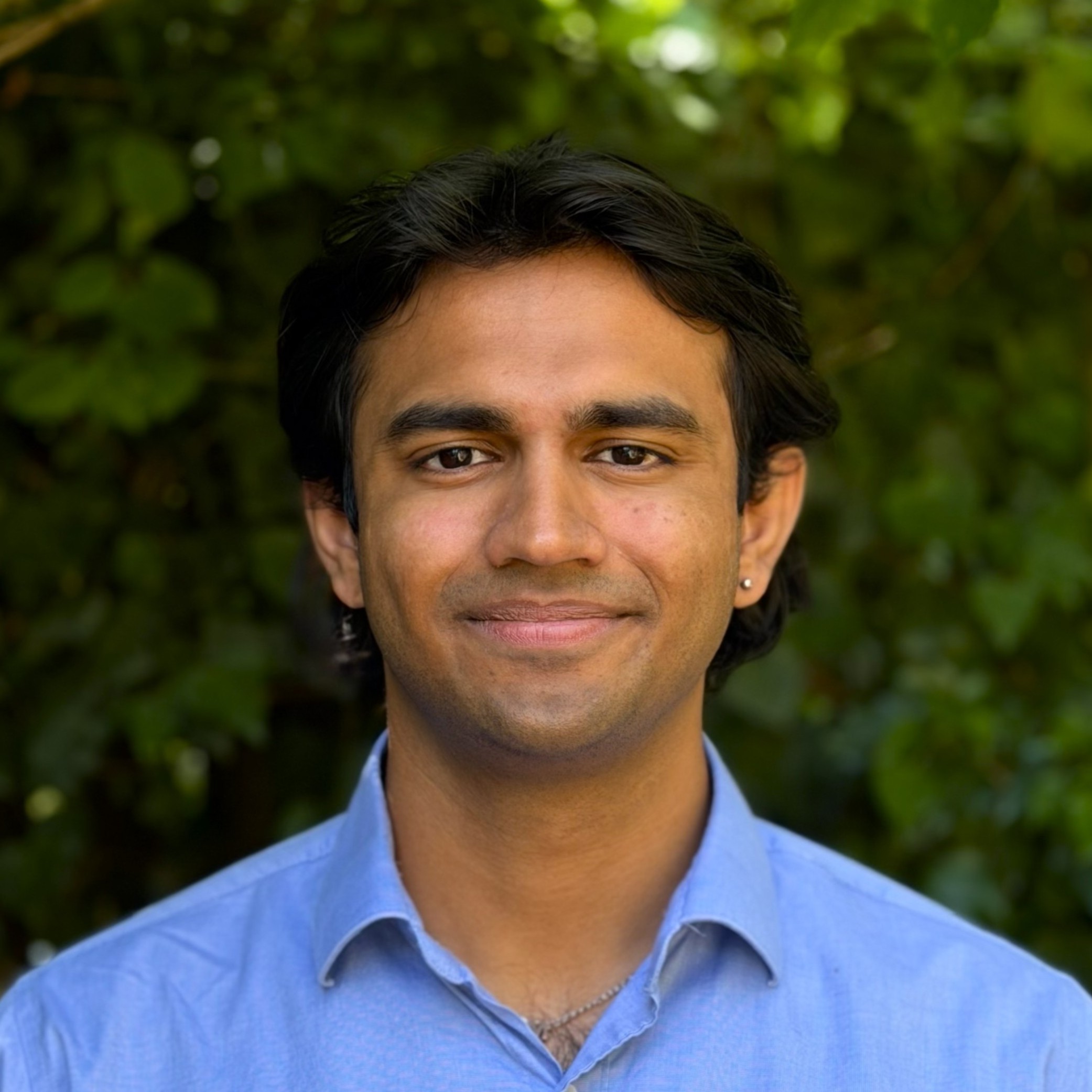 Close-up of a smiling man wearing a light blue shirt, outdoors with green foliage in the background.