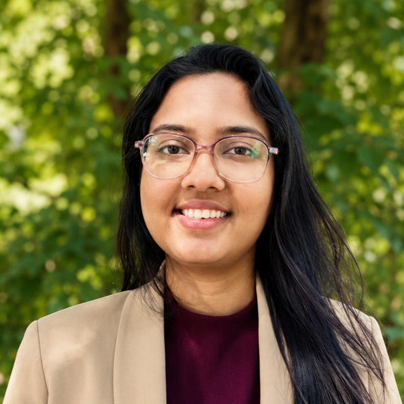 Woman with long dark hair and glasses smiling outdoors, wearing a beige blazer and burgundy top, greenery in the background.