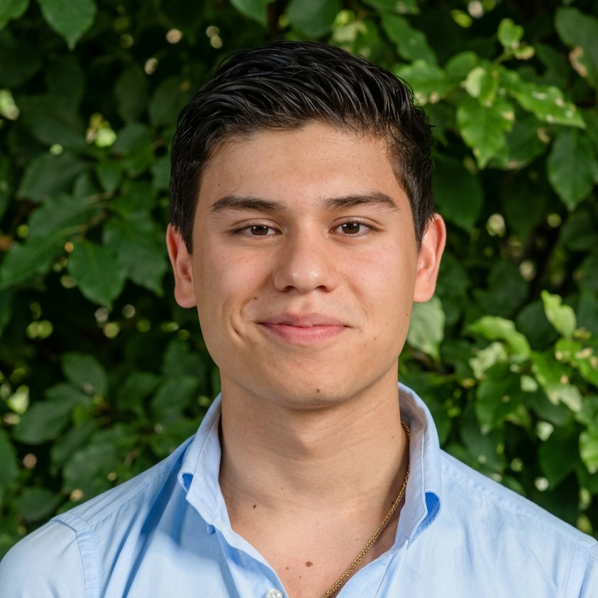 Head-and-shoulders portrait of a young man with short dark hair smiling, wearing a light blue button-down shirt outdoors with green foliage in the background.