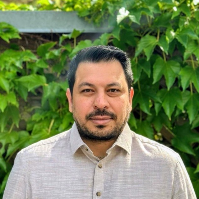 Man with short dark hair and beard wearing a light beige shirt, posing in front of green foliage.