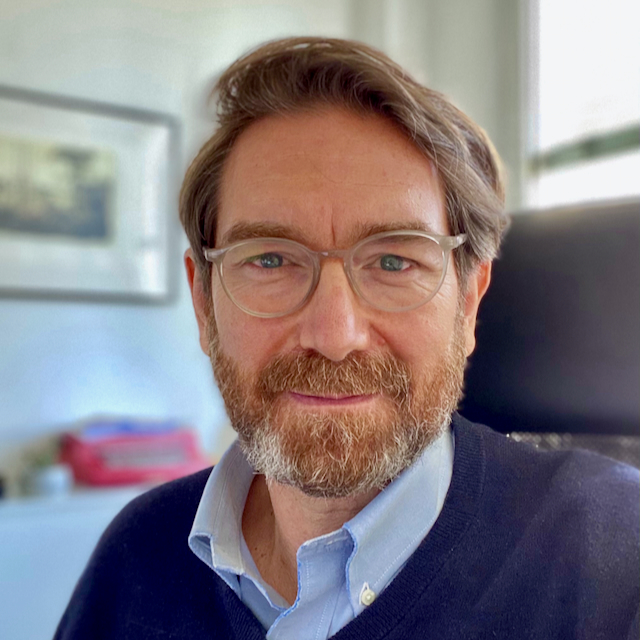 Friendly portrait of a man with light brown hair and beard, wearing glasses and a blue shirt in an office setting.